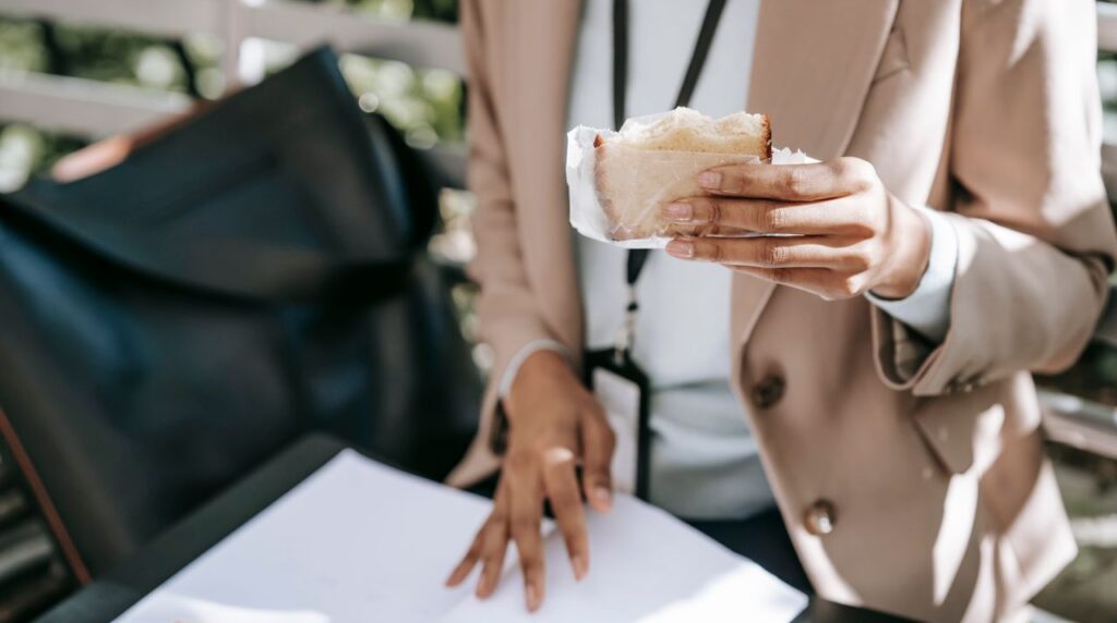 High angle of crop faceless African American female student reading documents and eating sandwich on bench in park